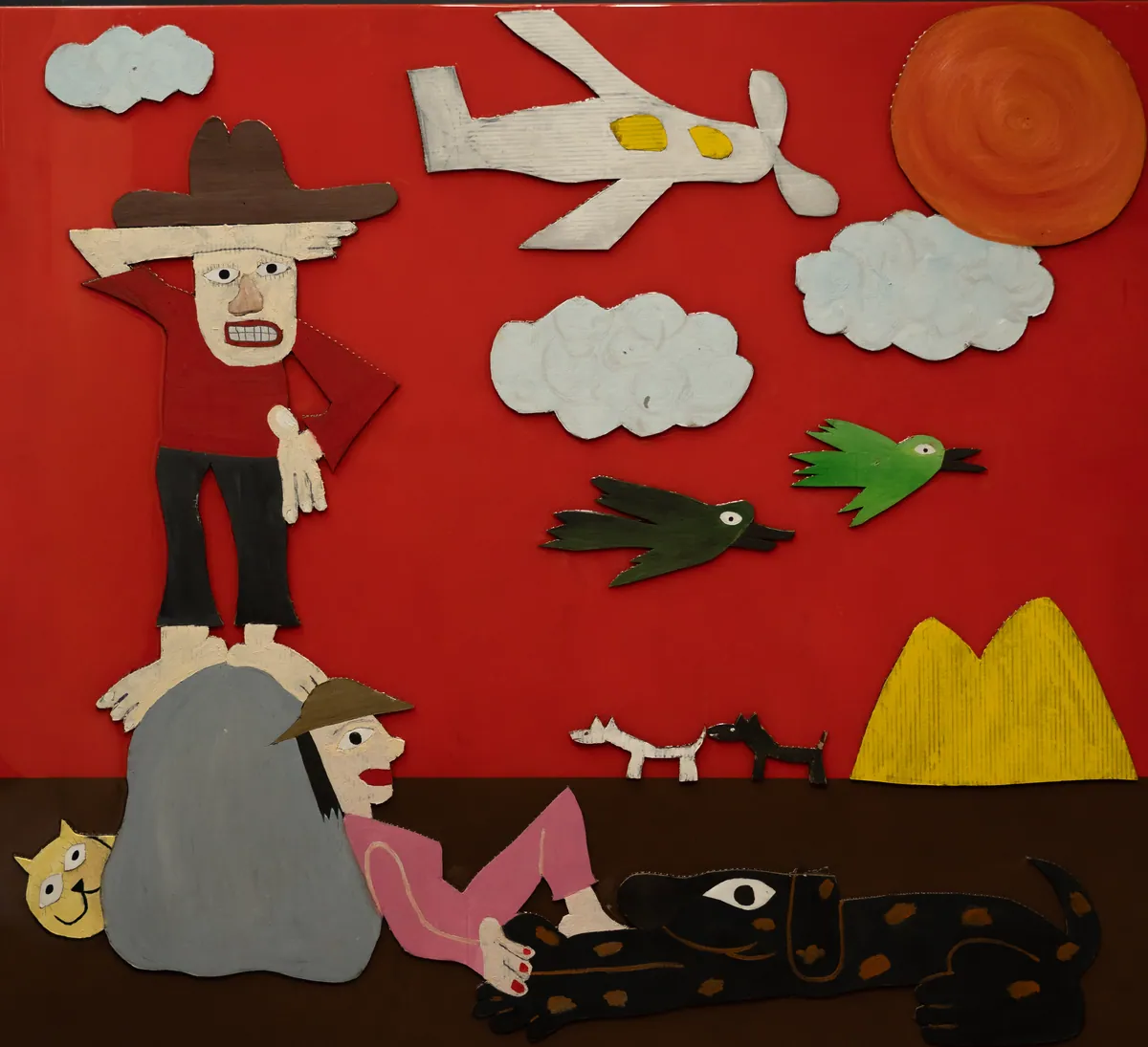 Full view: painted cardboard cut-outs of a cowboy, plane, birds and dogs on a glossy red ground