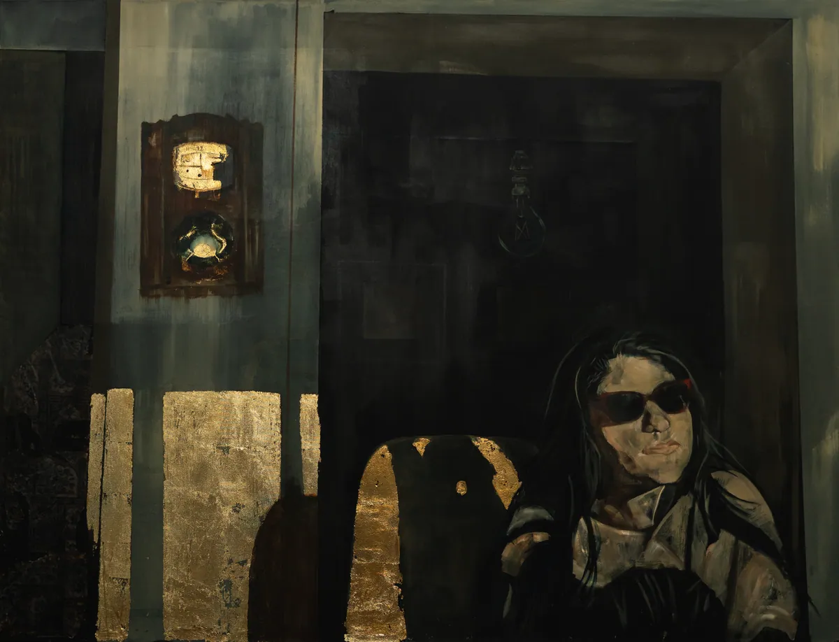 Full view: a seated figure in sunglasses in a dark interior, gold panels and a lit clock behind